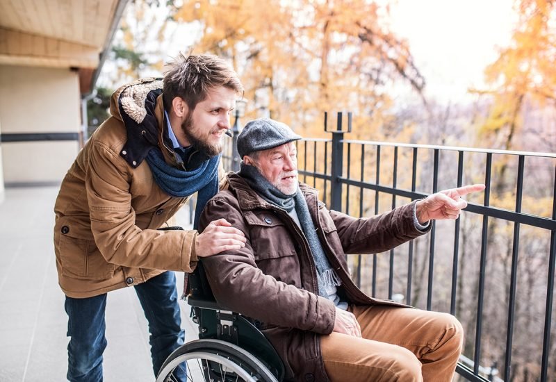 Father in wheelchair and young son on a walk. A carer assisting disabled senior man.